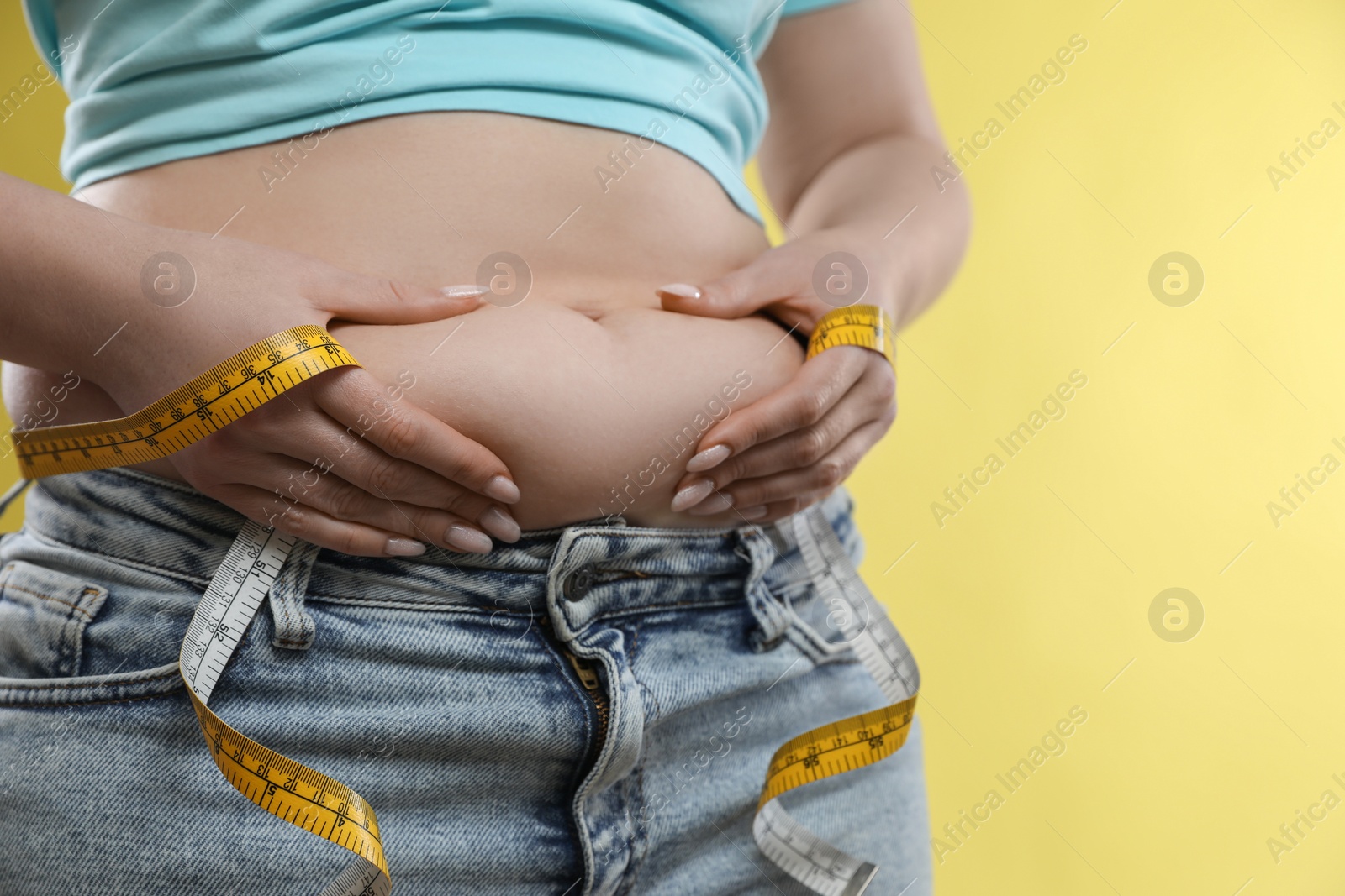 Photo of Woman with measuring tape touching belly fat on yellow background, closeup. Overweight problem