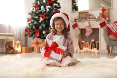 Photo of Cute little child in Santa hat with Christmas gift box at home