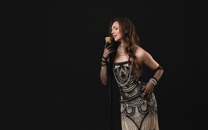 Beautiful young woman in stylish dress with microphone singing on black background, space for text