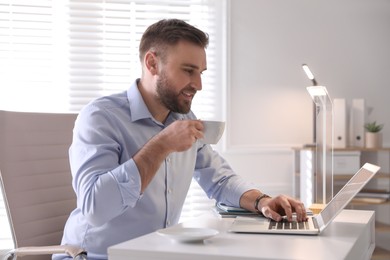 Photo of Young man drinking coffee while working on laptop at table in office