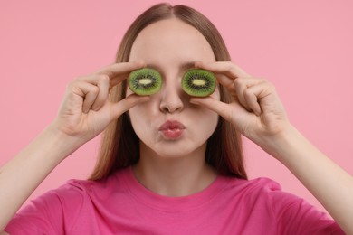 Young woman holding halves of kiwi near her eyes on pink background