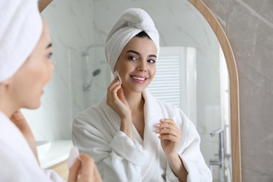Photo of Beautiful young woman cleaning her face with cotton disk near mirror in bathroom