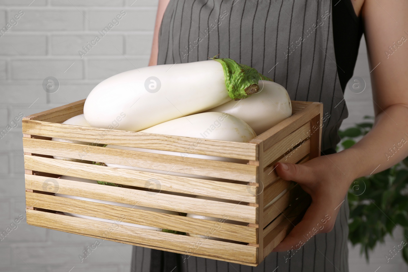 Photo of Woman holding wooden crate with white eggplants near brick wall, closeup