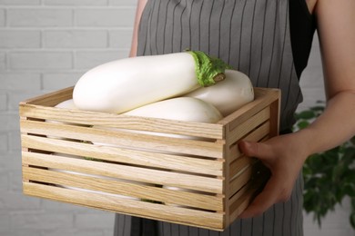 Woman holding wooden crate with white eggplants near brick wall, closeup