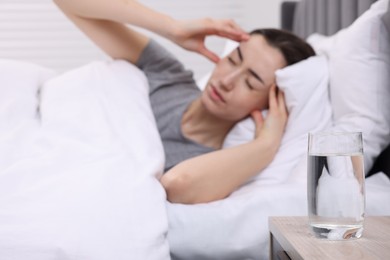 Sad woman suffering from headache in bed indoors, focus on glass of water