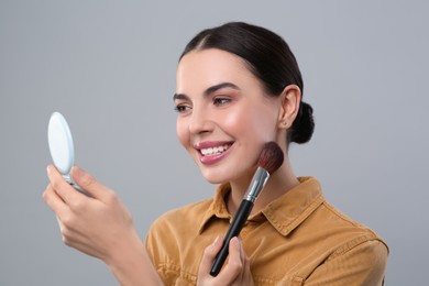 Happy woman with cosmetic pocket mirror applying makeup on light grey background