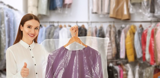 Dry-cleaning service. Happy woman holding hanger with sweatshirt in plastic bag and showing thumbs up indoors, space for text. Banner design
