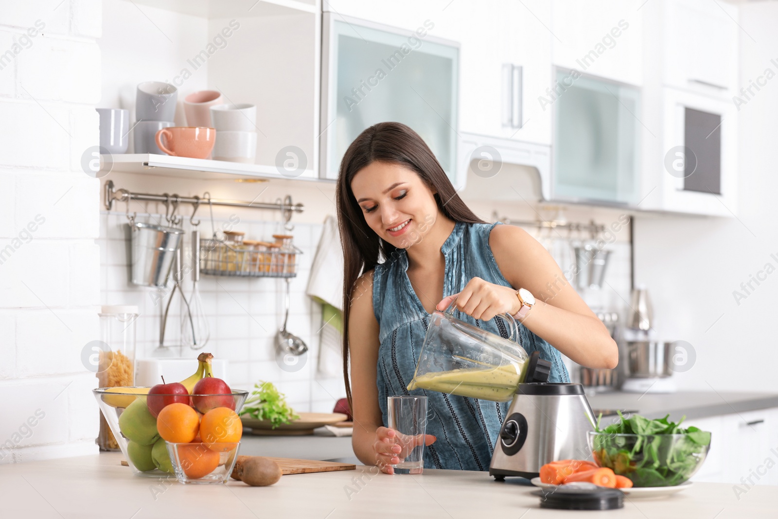 Photo of Young woman pouring tasty healthy smoothie into glass at table in kitchen