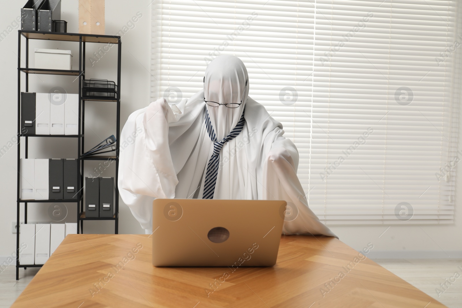 Photo of Overworked ghost. Man covered with white sheet using laptop at wooden table in office