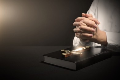 Woman holding hands clasped while praying at table with Bible and cross against dark background, closeup. Space for text