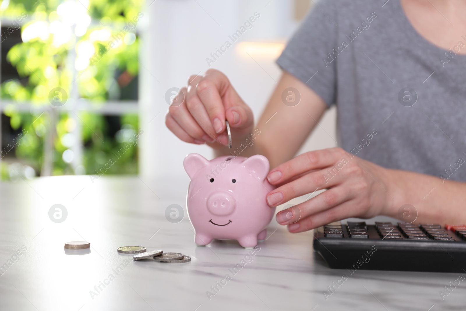 Photo of Woman putting money into piggy bank at marble table indoors, closeup