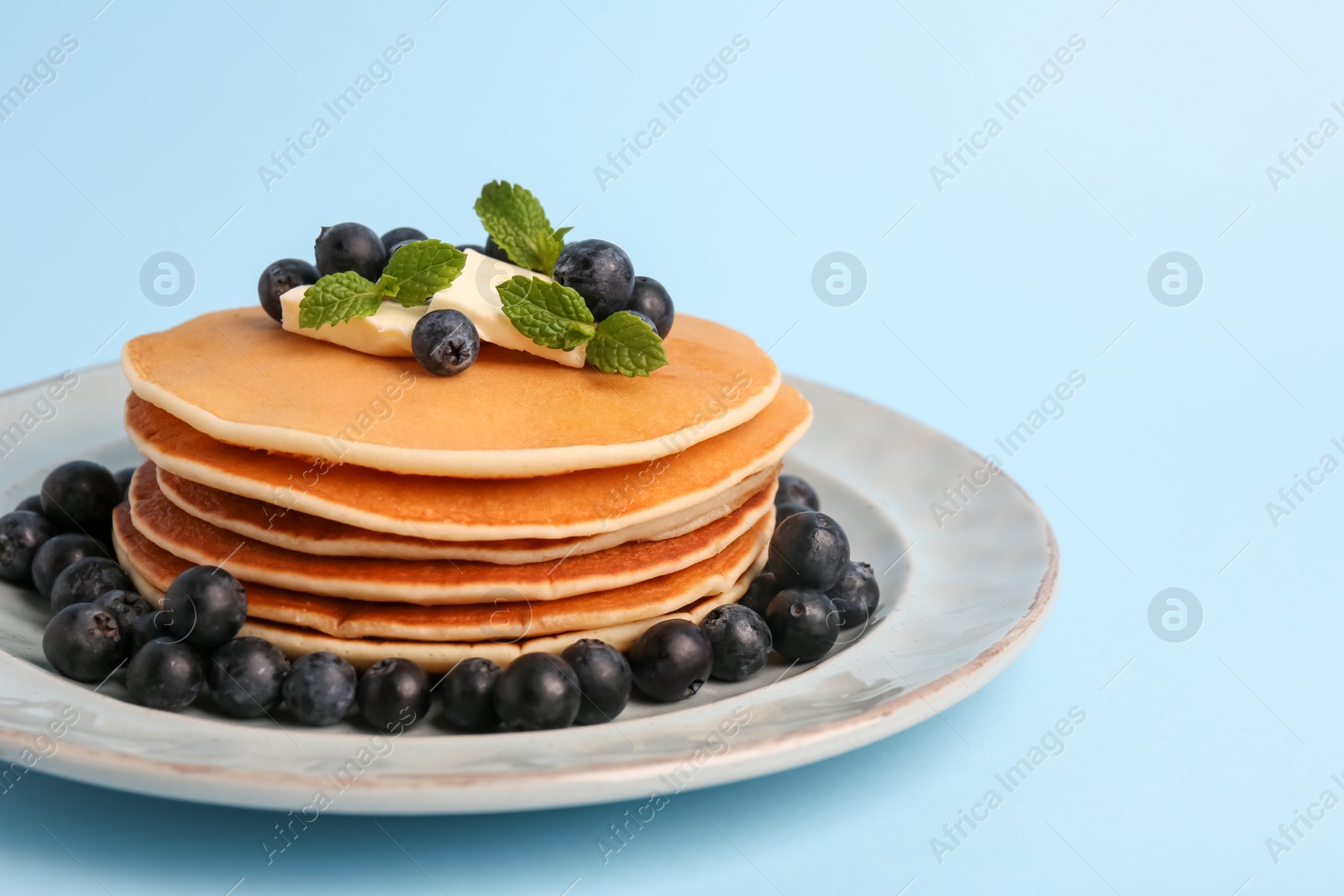 Photo of Stack of tasty pancakes with blueberries, butter and mint on light blue background, closeup. Space for text