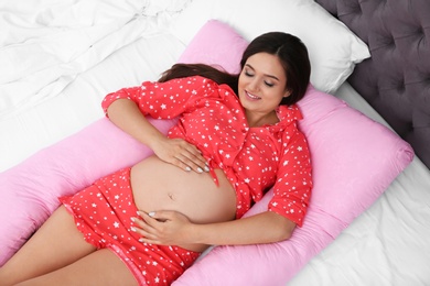 Beautiful pregnant woman lying with maternity pillow on bed at home, top view