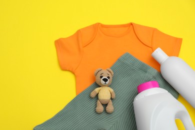 Bottles of laundry detergents, baby clothes and toy bear on yellow background, flat lay