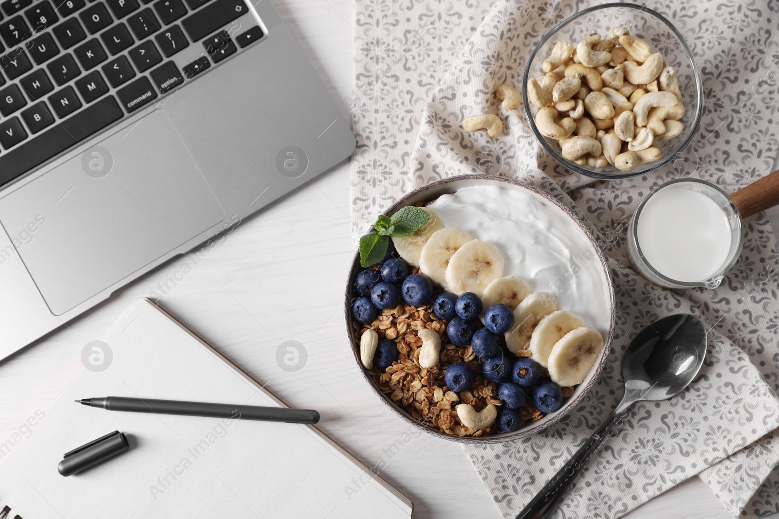 Photo of Delicious granola in bowl, stationery and laptop on white wooden table, flat lay