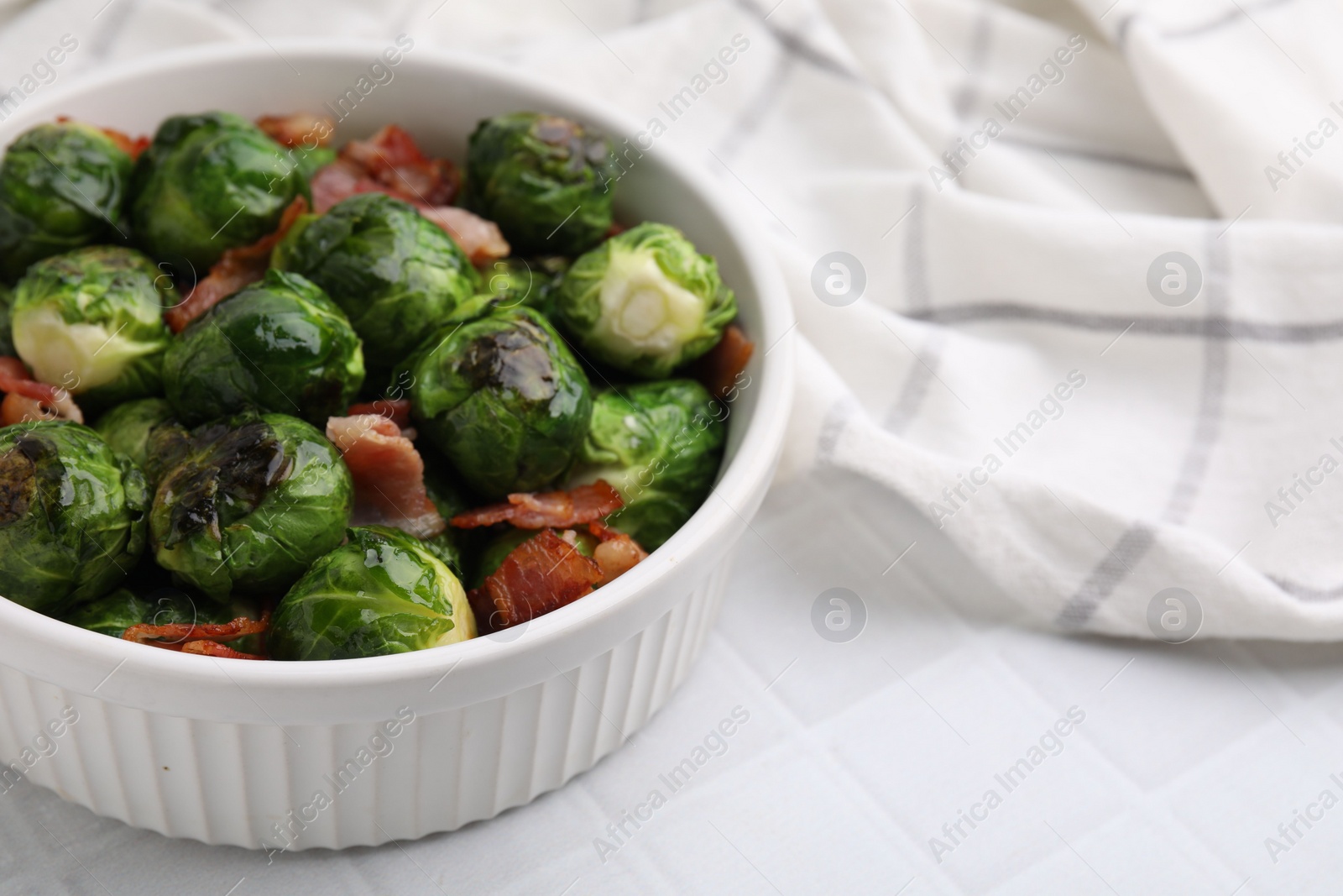 Photo of Delicious roasted Brussels sprouts and bacon in bowl on white tiled table, closeup. Space for text