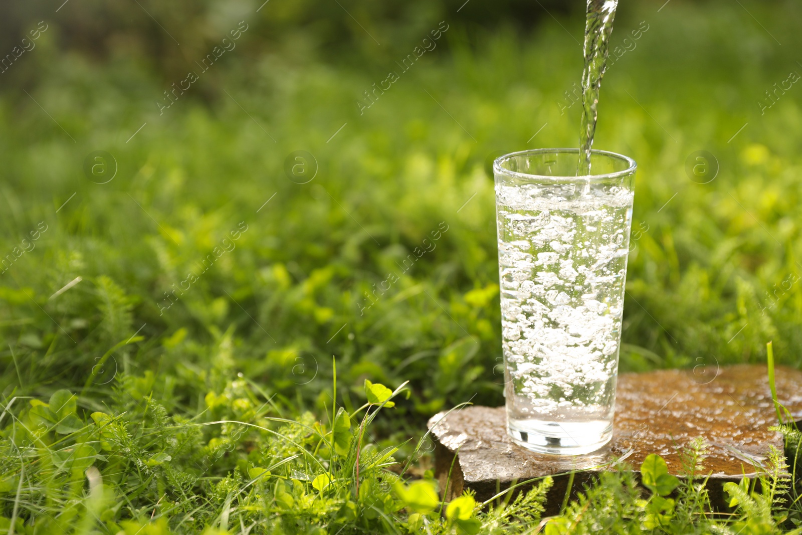 Photo of Pouring fresh water into glass on stone in green grass outdoors. Space for text