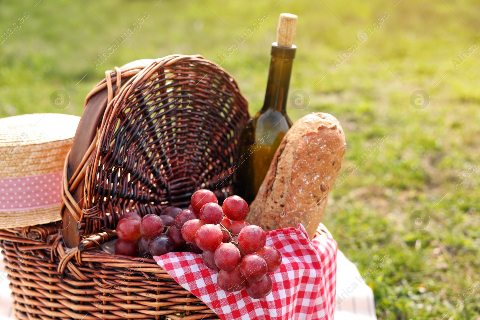 Photo of Wicker picnic basket with bottle of wine, bread, grapes and napkin on green grass outdoors, closeup. Space for text