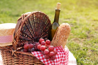 Photo of Wicker picnic basket with bottle of wine, bread, grapes and napkin on green grass outdoors, closeup. Space for text