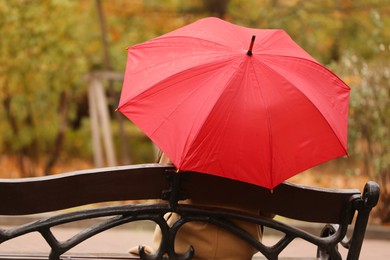 Photo of Woman with red umbrella sitting on bench in autumn park, back view