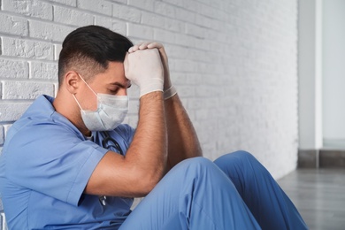 Photo of Sad doctor sitting near white brick wall indoors. Stress of health care workers during coronavirus pandemic