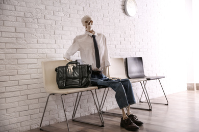 Photo of Human skeleton in office wear sitting on chair near brick wall indoors