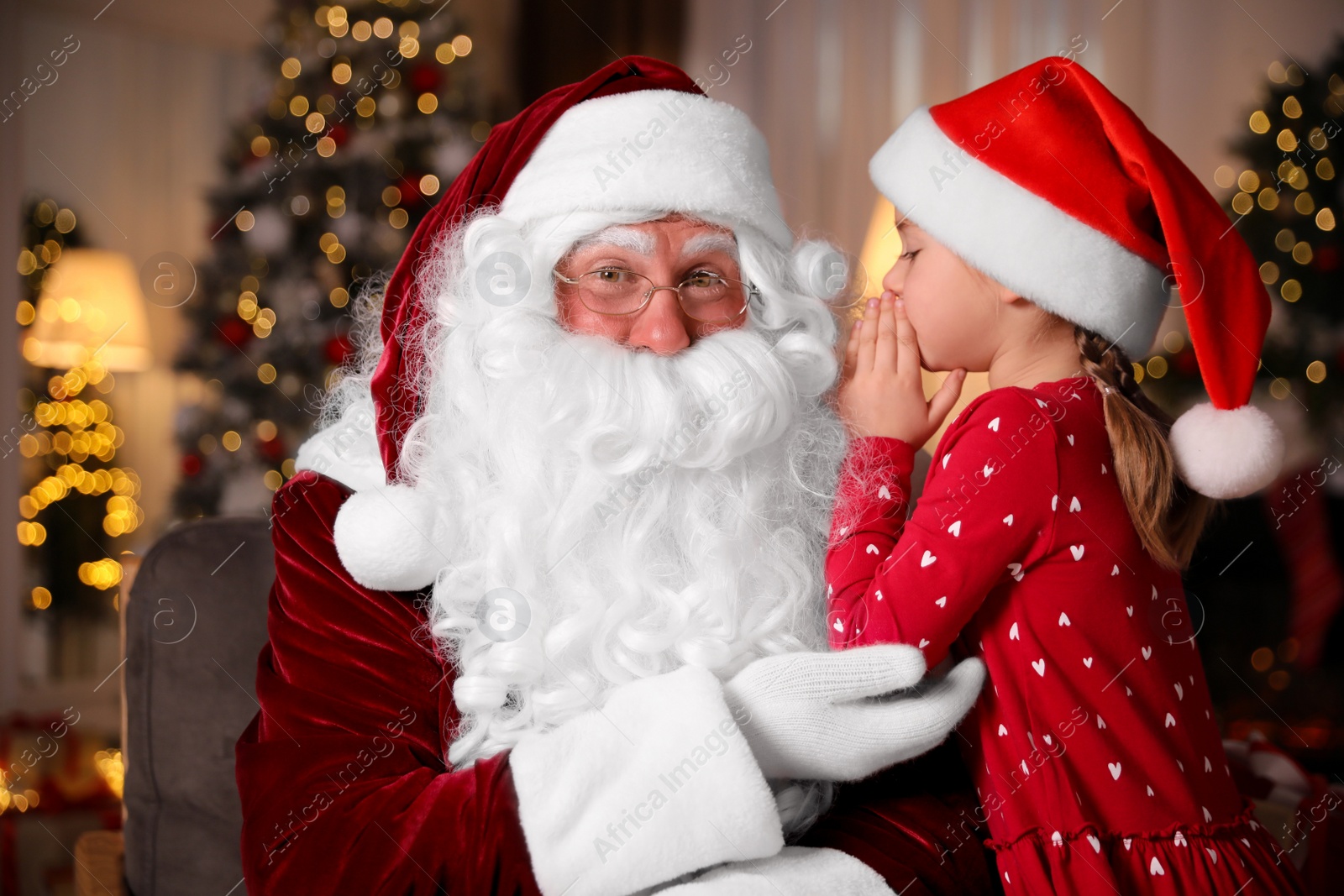 Photo of Little girl whispering in Santa Claus' ear near Christmas tree indoors