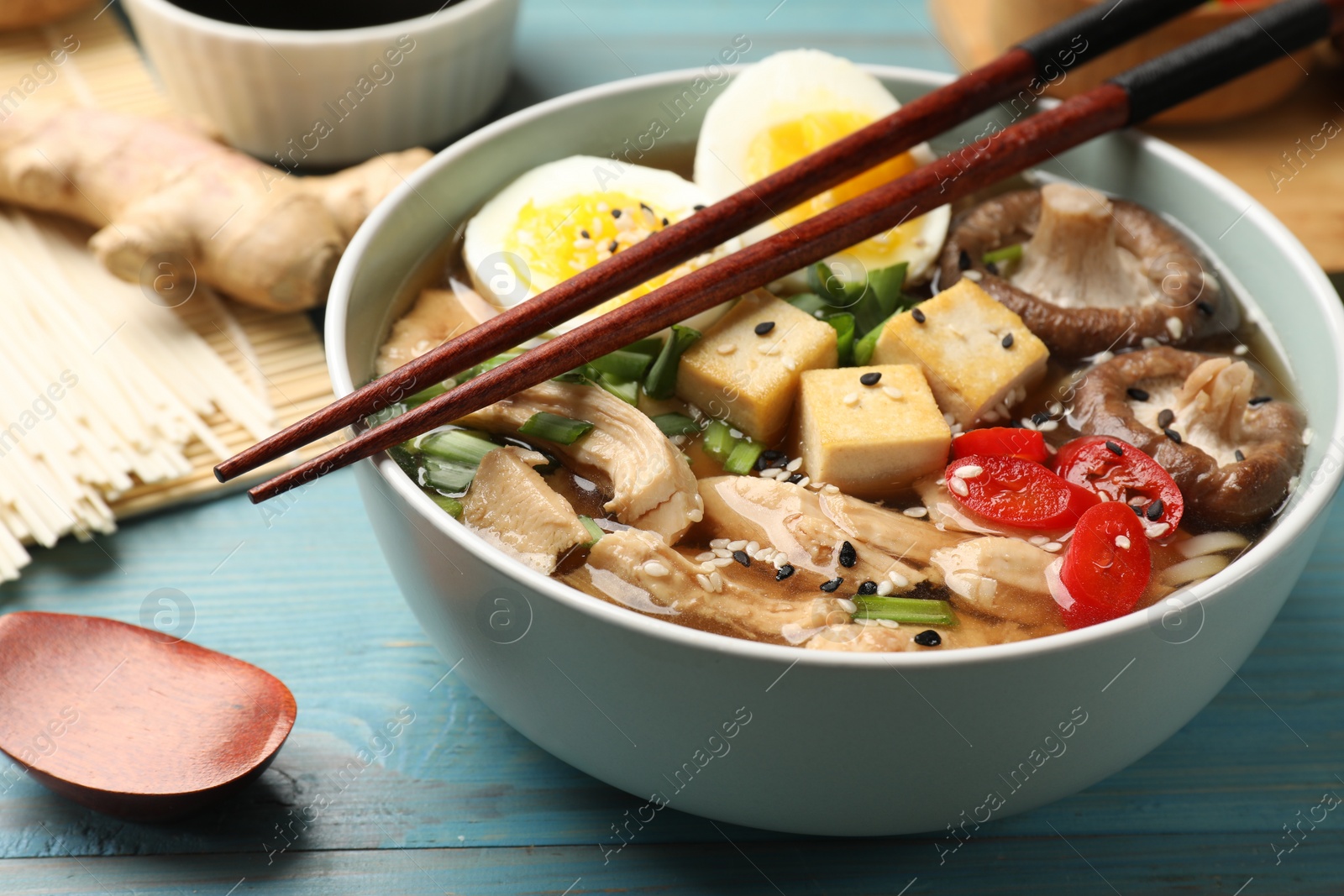 Photo of Bowl of delicious ramen and ingredients on light blue wooden table, closeup. Noodle soup