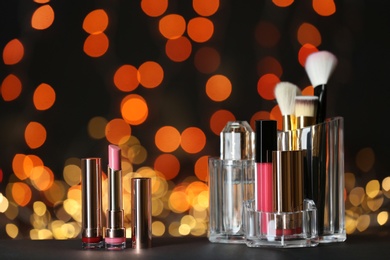 Photo of Bright lipsticks and holder with other makeup products on table against blurred lights