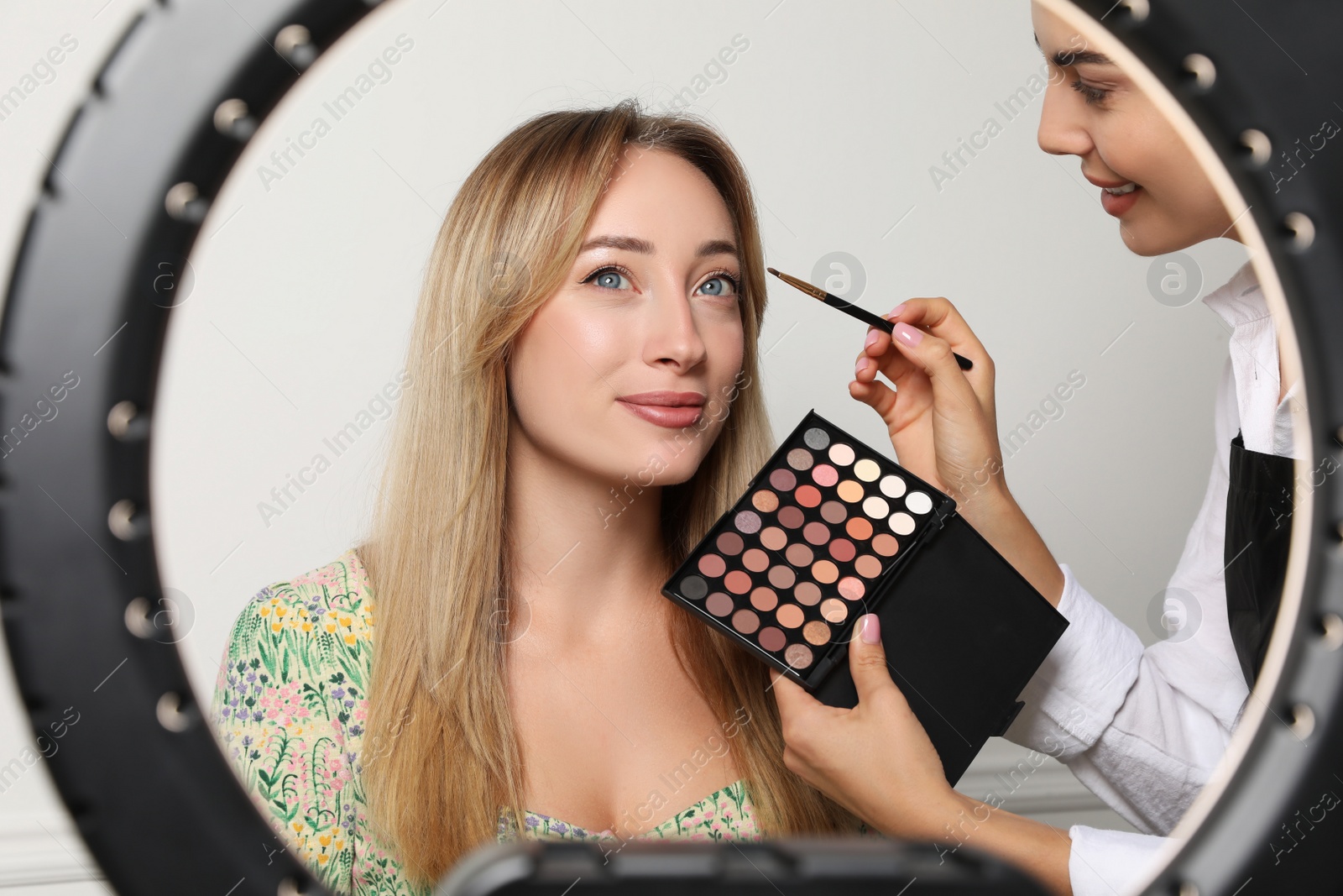 Photo of Professional makeup artist working with beautiful young woman on light background, view through ring lamp