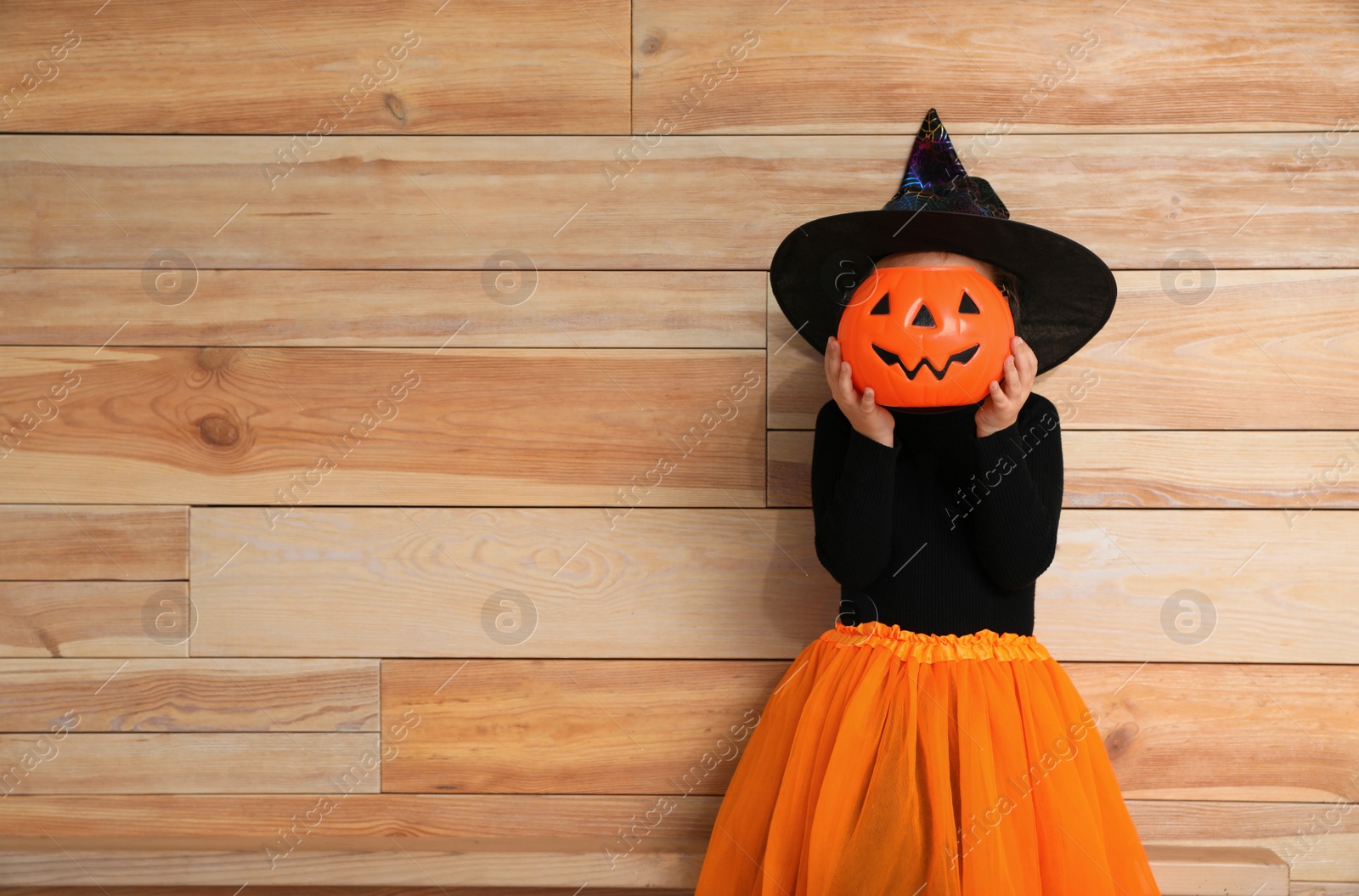 Photo of Cute little girl with pumpkin candy bucket wearing Halloween costume on wooden background. Space for text