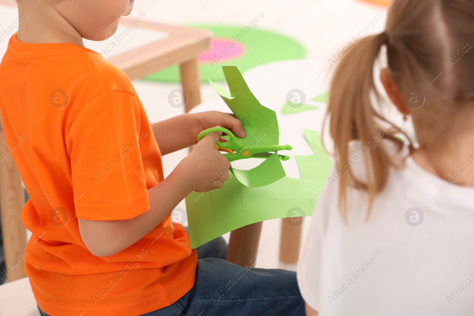Photo of Little girl and boy cutting color paper with scissors at desk, closeup. Kindergarten activities