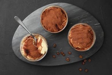 Photo of Delicious tiramisu in glasses, spoon and coffee beans on black table, top view