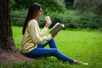 Photo of Young woman with cup of coffee reading book near tree in park