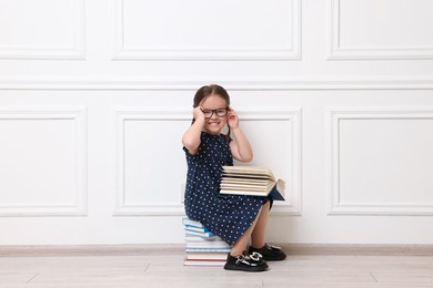 Photo of Cute little girl in glasses sitting on stack of books near white wall