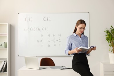 Young chemistry teacher reading book near whiteboard in classroom. Space for text