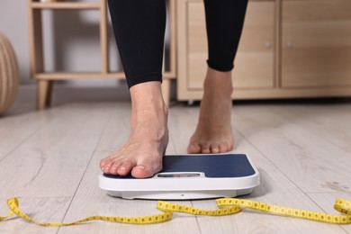 Photo of Woman stepping on floor scale and measuring tape at home, closeup. Weight control