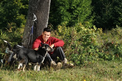 Photo of Hiker stroking dog while sitting near tree on grass. Rest stop
