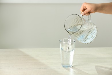 Woman pouring water from jug into glass on white table indoors, closeup. Space for text