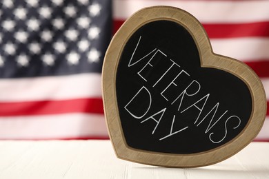 Photo of Heart shaped blackboard with phrase Veterans Day on white wooden table against American flag, space for text