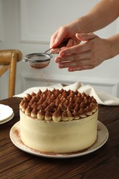 Photo of Woman dusting delicious tiramisu cake with cocoa powder at wooden table, closeup