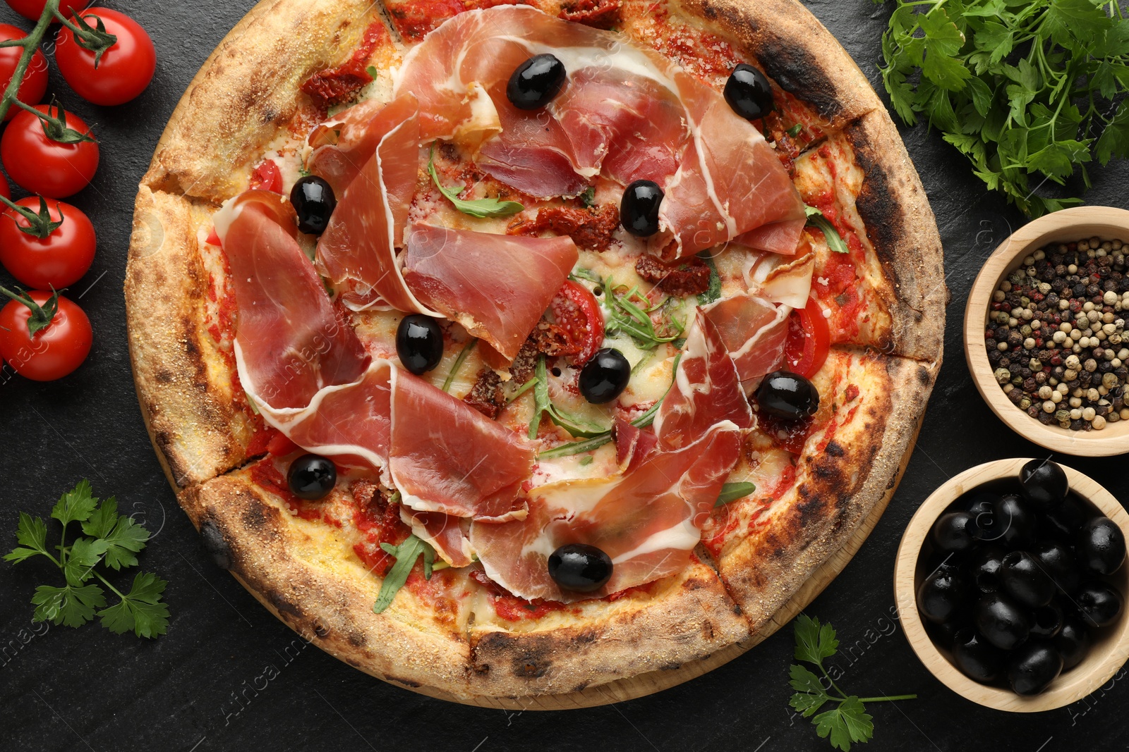 Photo of Tasty pizza with cured ham, olives, tomatoes, parsley and spices on black table, top view