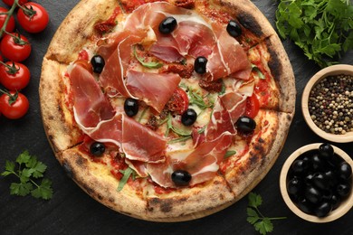 Tasty pizza with cured ham, olives, tomatoes, parsley and spices on black table, top view