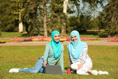 Muslim women in hijabs with laptop sitting on green lawn outdoors