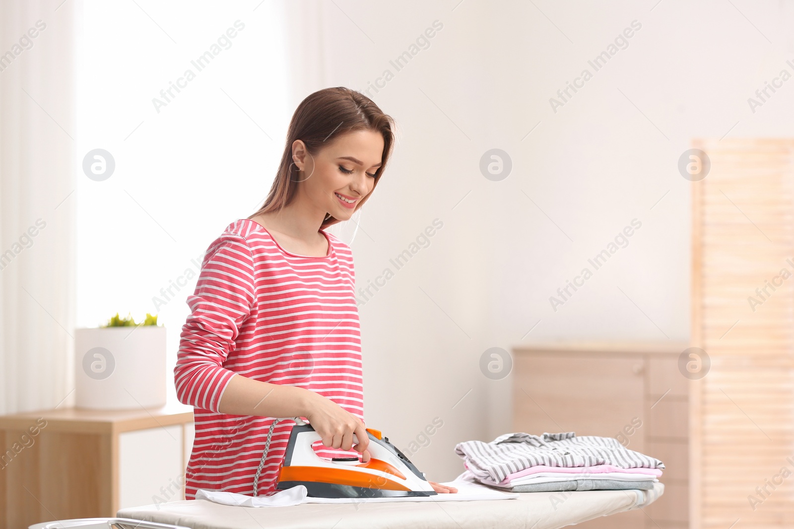 Photo of Young pretty woman ironing clean laundry indoors. Space for text