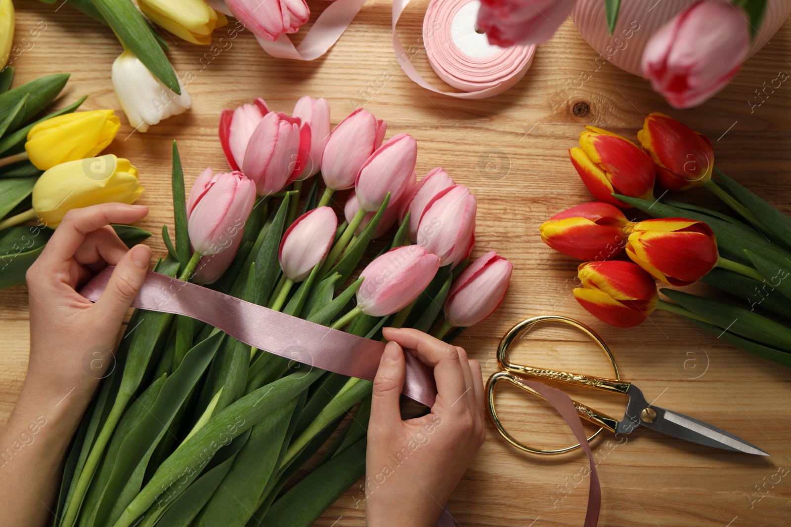 Photo of Woman making beautiful bouquet of fresh tulips and ribbon at wooden table, top view