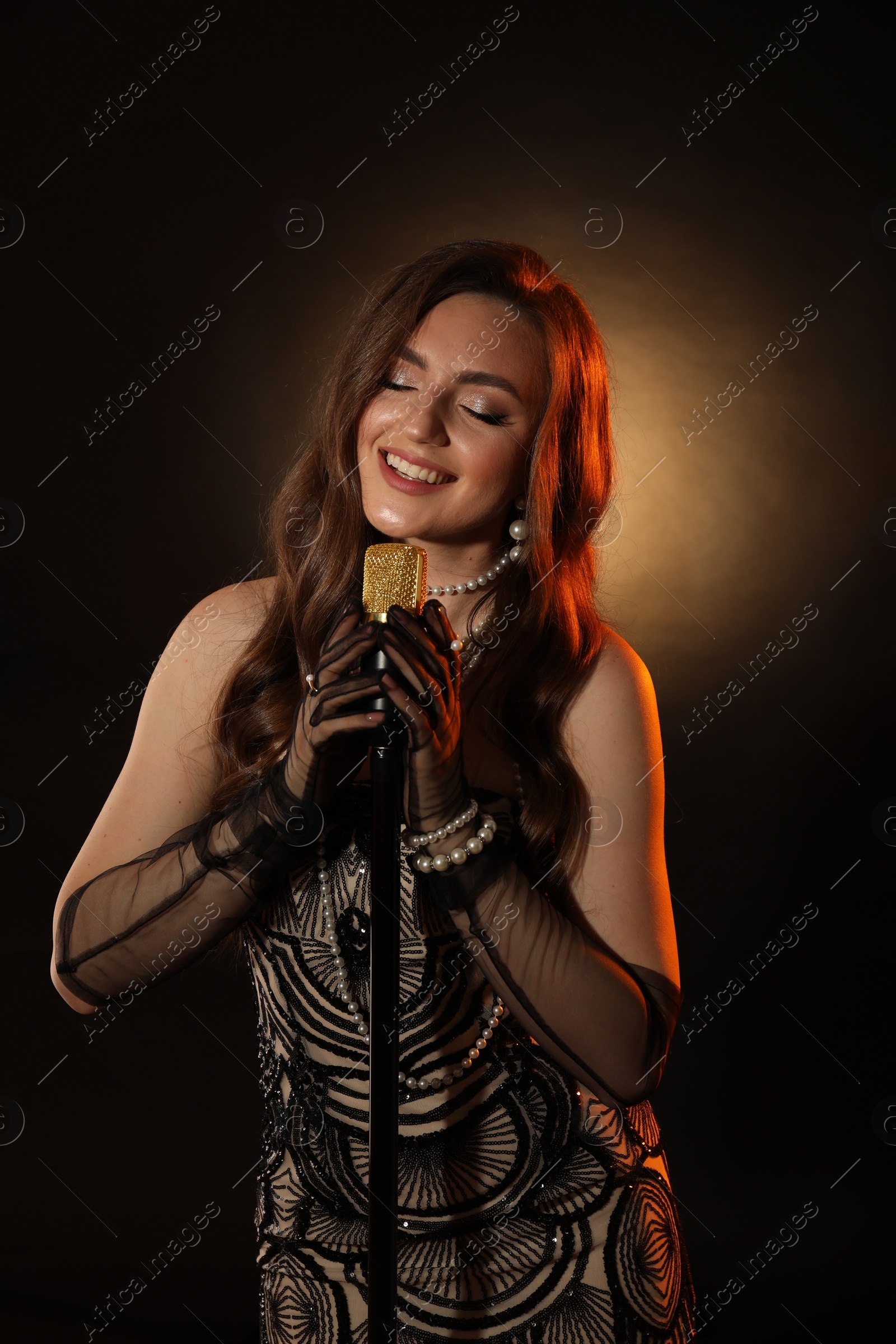 Photo of Beautiful young woman in stylish dress with microphone singing on dark background