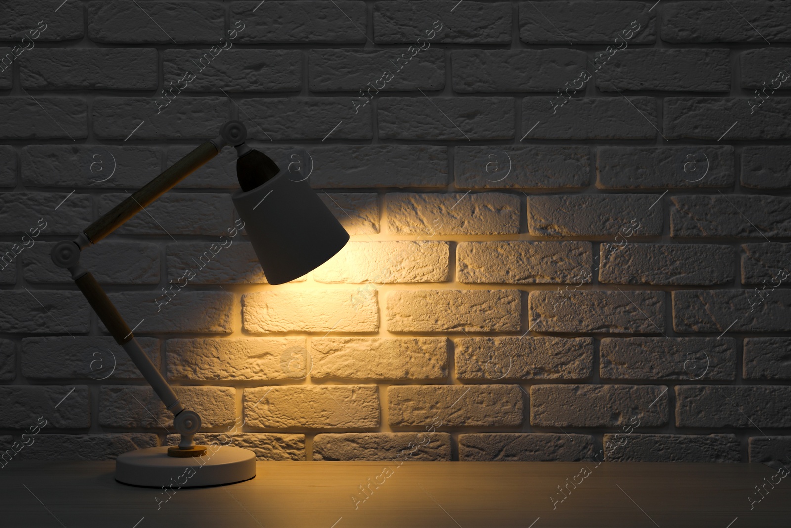 Photo of Stylish modern desk lamp on white wooden table near brick wall at night, space for text