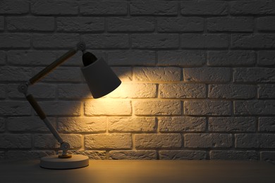 Photo of Stylish modern desk lamp on white wooden table near brick wall at night, space for text