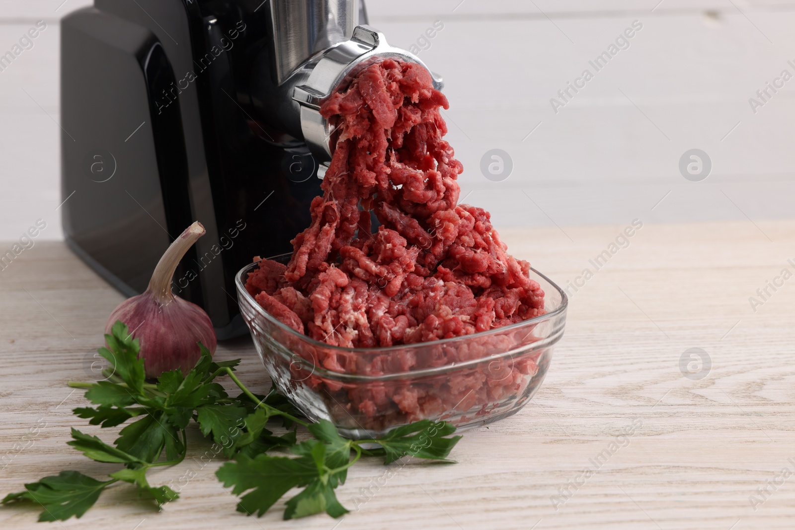 Photo of Electric meat grinder with beef mince, garlic and parsley on white wooden table, space for text